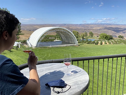 Side view of Andrew tasting wine on a patio with a green lawn, domed concert venue, and Columbia River gorge behind him