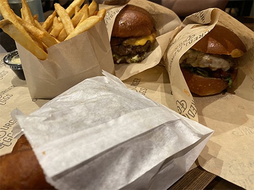 Fries on the left, two burgers in brown wax paper on the right, and a bag of onion rings in front