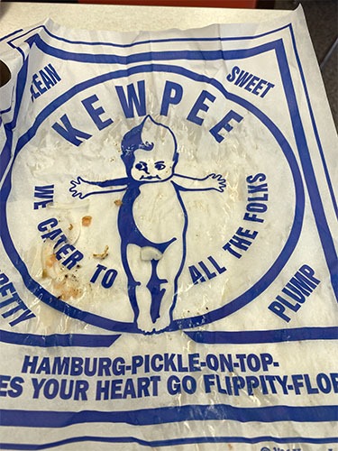 Kewpee burger wrapper with a picture of the doll. The text says "Kewpee - We cater to all  the folks" and "Hamburg-pickle-on-top makes your heart go flippity flop"