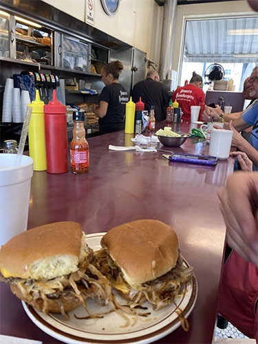 Two small burgers with piles of onions coming out from under the bun, sitting on a plate, on a diner counter. The restaurant staff is busy in the background.