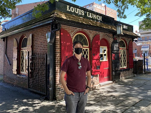 Andrew standing outside a very small, old brick building with the sign Louis' Lunch and  red door.
