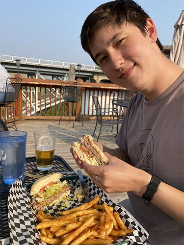 Andrew holding half a cut burger, the other half in a basket with fries, sitting outside on a deck