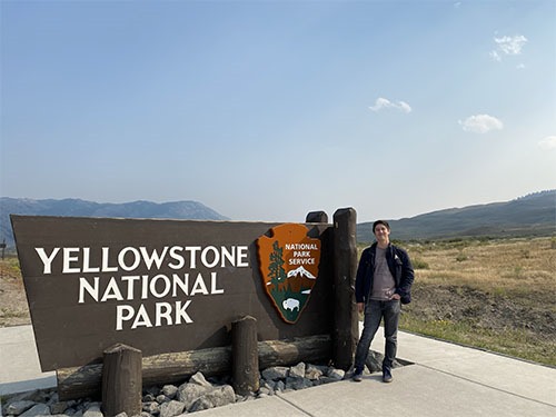 Andrew standing next to a large wooden sign saying Yellowstone National Park