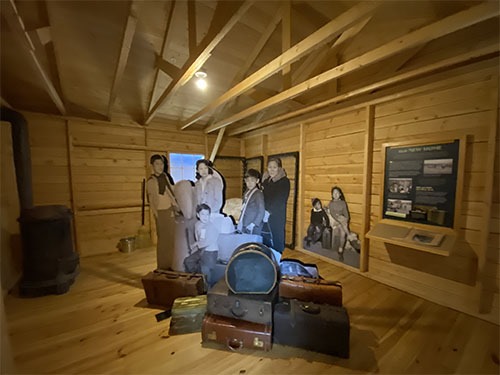 A pile of luggage and lifesize photo cutouts of a Japanese American family in the middle of an empty, bare wood barracks.
