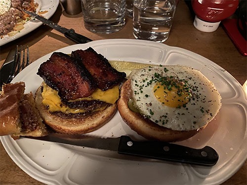 Open burger on a plate with two thick strips of bacon topping one side and a fried egg topping the other side.