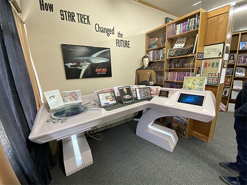 A Star Fleet desk with a cardboard cutout of Data behind and other pictures and books on top of it.