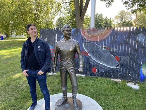 Andrew standing next to a near life-size bronze sculpture of Captain Kirk. Behind is a wood fence with a Star Trek mural