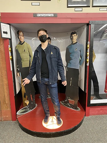 Andrew standing in an original series-like transporter bay in front of cardboard cutouts of Kirk, McCoy, and Spock.