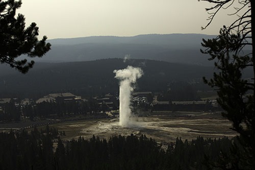 A geyser erupting high into the air as seen from a high vantage. A big lodge is beyind the geyser