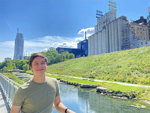 Andrew standing on a walkway in front of an old canal with a set of tall concrete grain silos behind him. A big sign saying "Gold Medal Flour" is on top of the silos