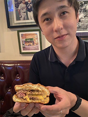 Andrew sitting in a restaurant booth, holding a burger cut in half to show a patty with a pocket inside and melted cheese oozing out.