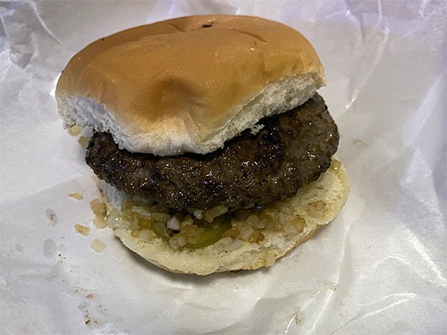 A thick hamburger on a paper wrapper. The patty sits on finely diced grilled onions and a pickle chip.