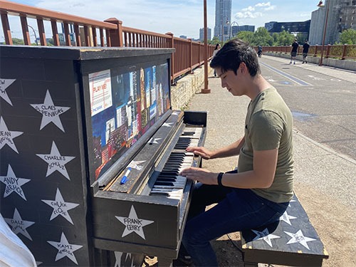 Andrew playing an upright piano painted with white stars and a mural. The piano is outside by a railing on the sidewalk.