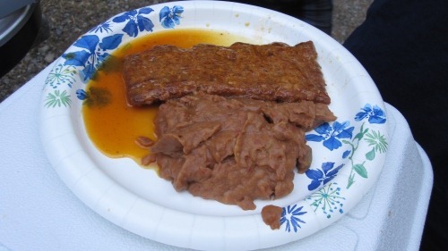A paper plate with refried beans and a slab of meat with some sauce/juices pooling.